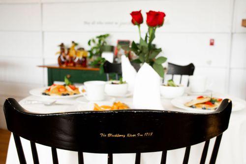 Breakfast table with chair in foreground. The back side of the chair is visible and has "The Blackbery River Inn 1763" written on it in golden letters.